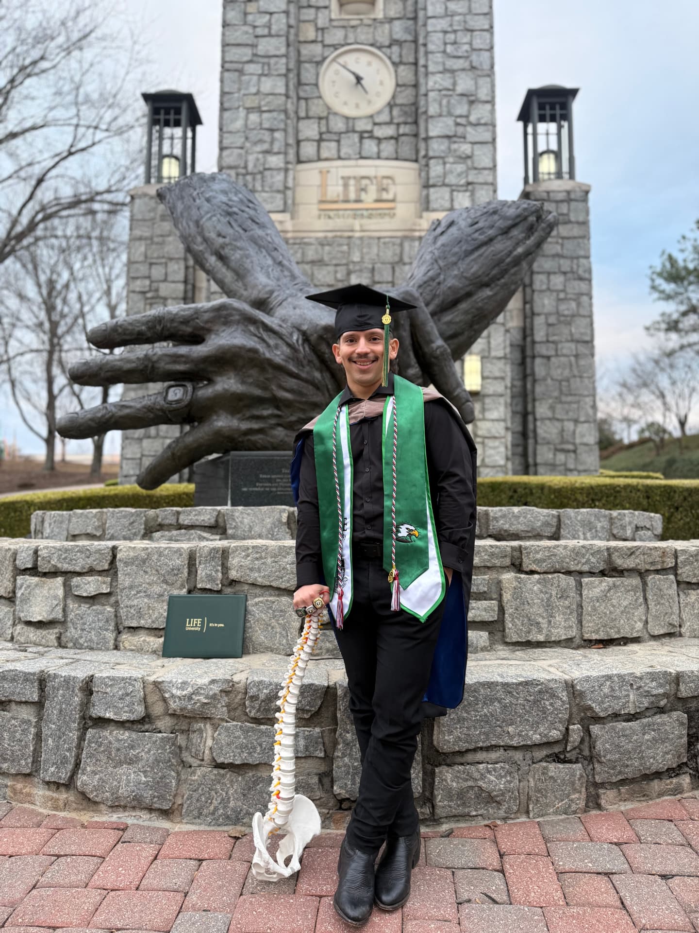 Dr. Adrian Escobedo in graduation regalia at Life University, holding a model spine